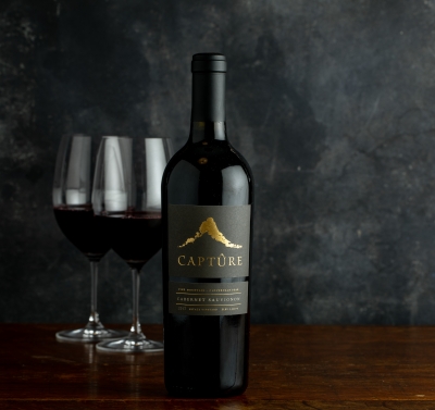 Single bottle of red wine against a dark background with two wine glasses on the left.