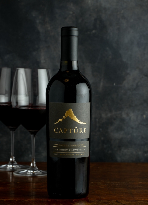 Single bottle of red wine against a dark background with two wine glasses on the left.