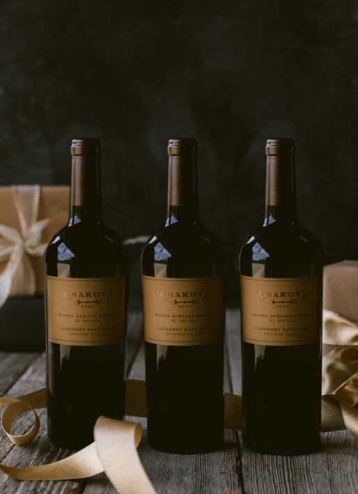 Three bottles of wine against a dark background with gifts behind the wines