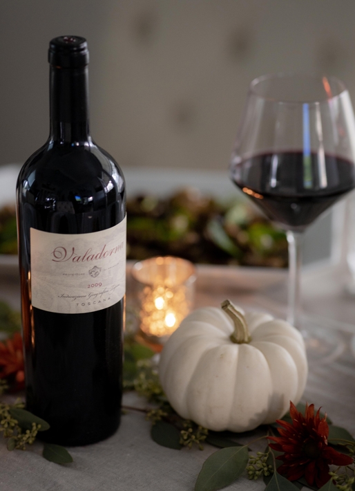 Bottle of wine, glass, and small white pumpkin on a table with holiday setting in the background.