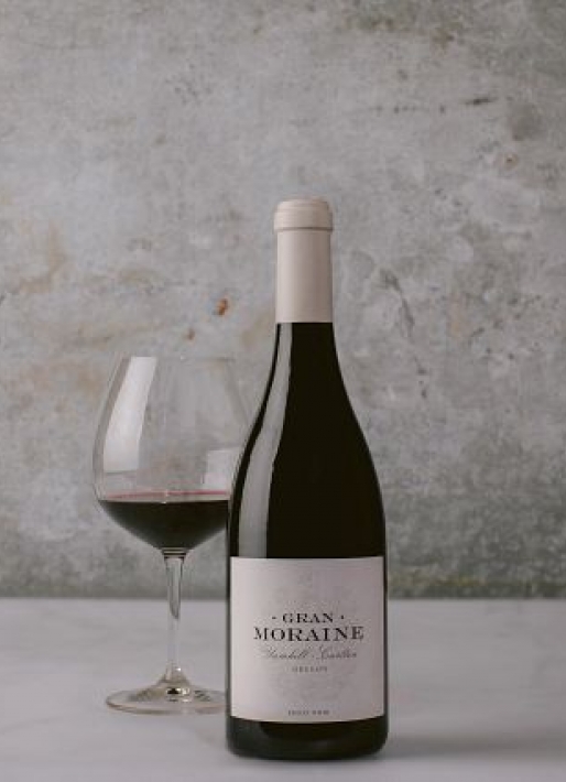 Single bottle of red wine against a dark background with one wine glass on the left.